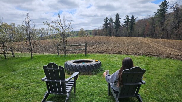 Two Adirondack chairs facing a circular fire pit in a grassy outdoor area with a fence and trees in the background. - Quaint 3-bedroom farmhouse in peaceful Mason, NH. (Mason)