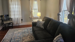 A living room with a dark-colored sofa, a patterned rug, a wooden chair, and a window with curtains. - Quaint 3-bedroom farmhouse in peaceful Mason, NH. (Mason)
