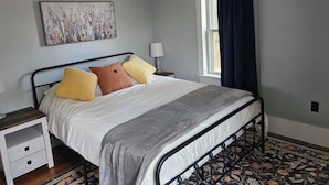 A bedroom with a bed, a nightstand, a lamp, a window with curtains, and a painting on the wall. - Quaint 3-bedroom farmhouse in peaceful Mason, NH. (Mason)