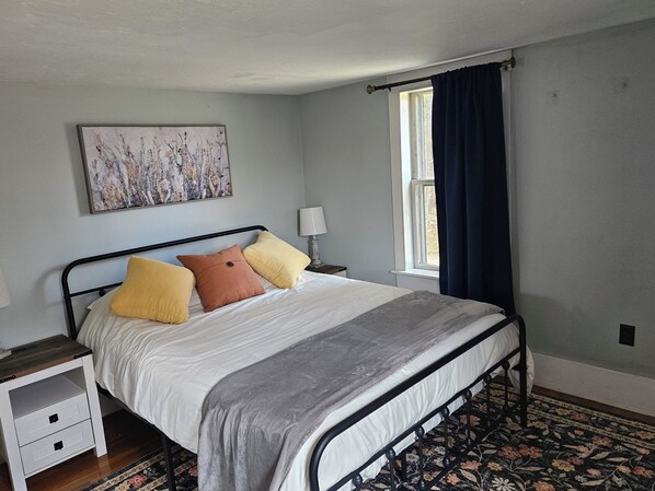 A bedroom with a bed, a nightstand, a lamp, a window with curtains, and a painting on the wall.