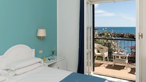 Ein Schlafzimmer mit Bett, Nachttisch, Lampe und Blick auf das Meer durch eine offene Tür.