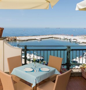 En terrasse med et bord dækket til to, med udsigt over en lysthavn med både og krystalklart hav.