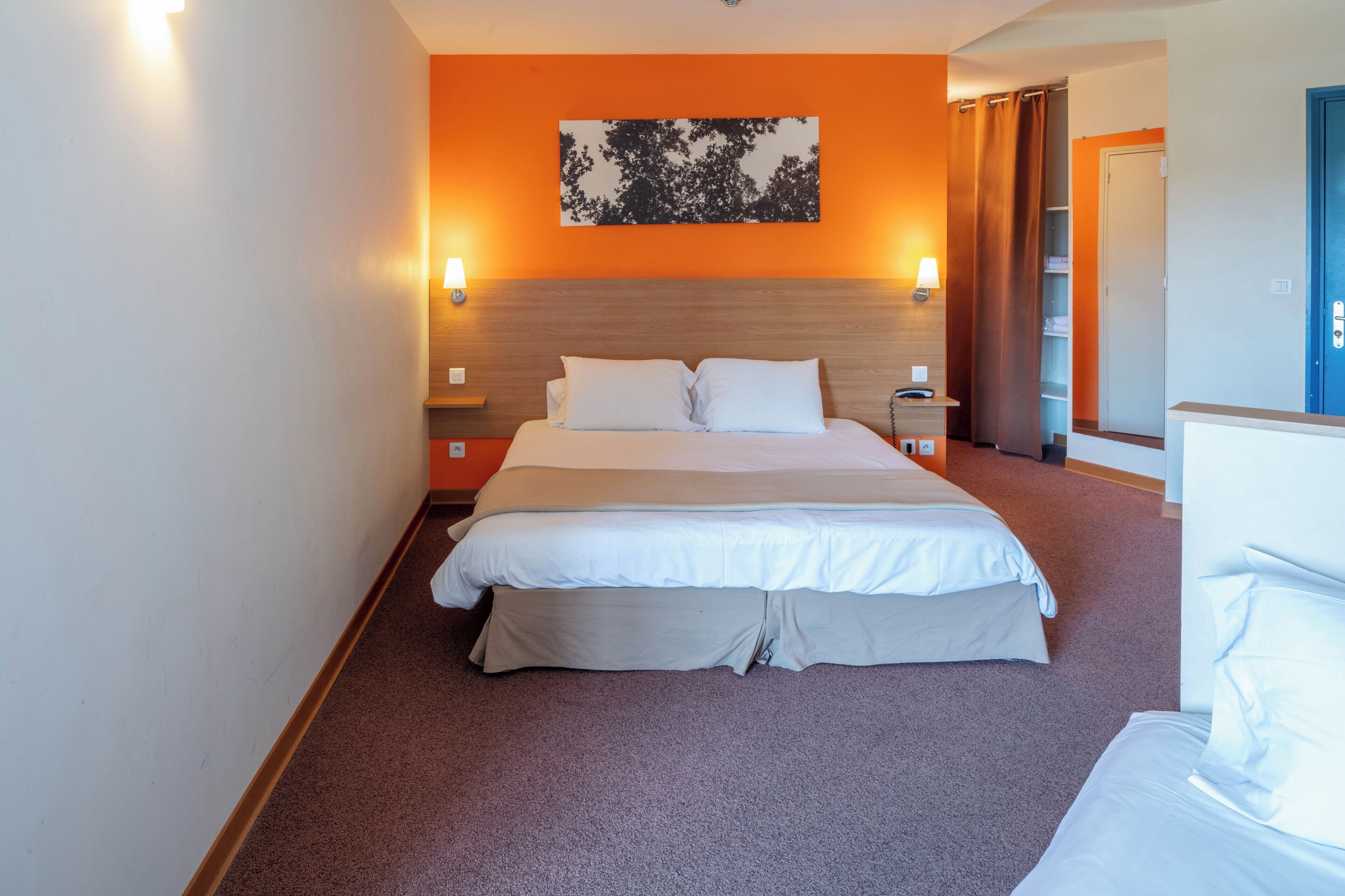 Une chambre d’hôtel avec un grand lit, deux tables de chevet, une petite table d’appoint, une lampe fixée au mur et un mur d’accent de couleur orange.