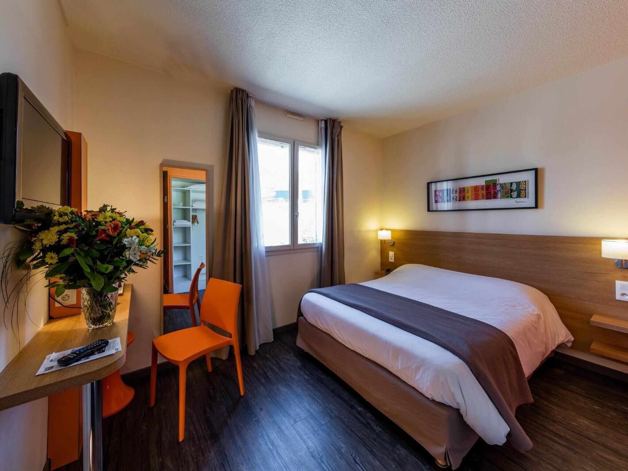 A hotel room with a bed, a desk with a chair, a TV, a window with curtains, and a framed picture on the wall.