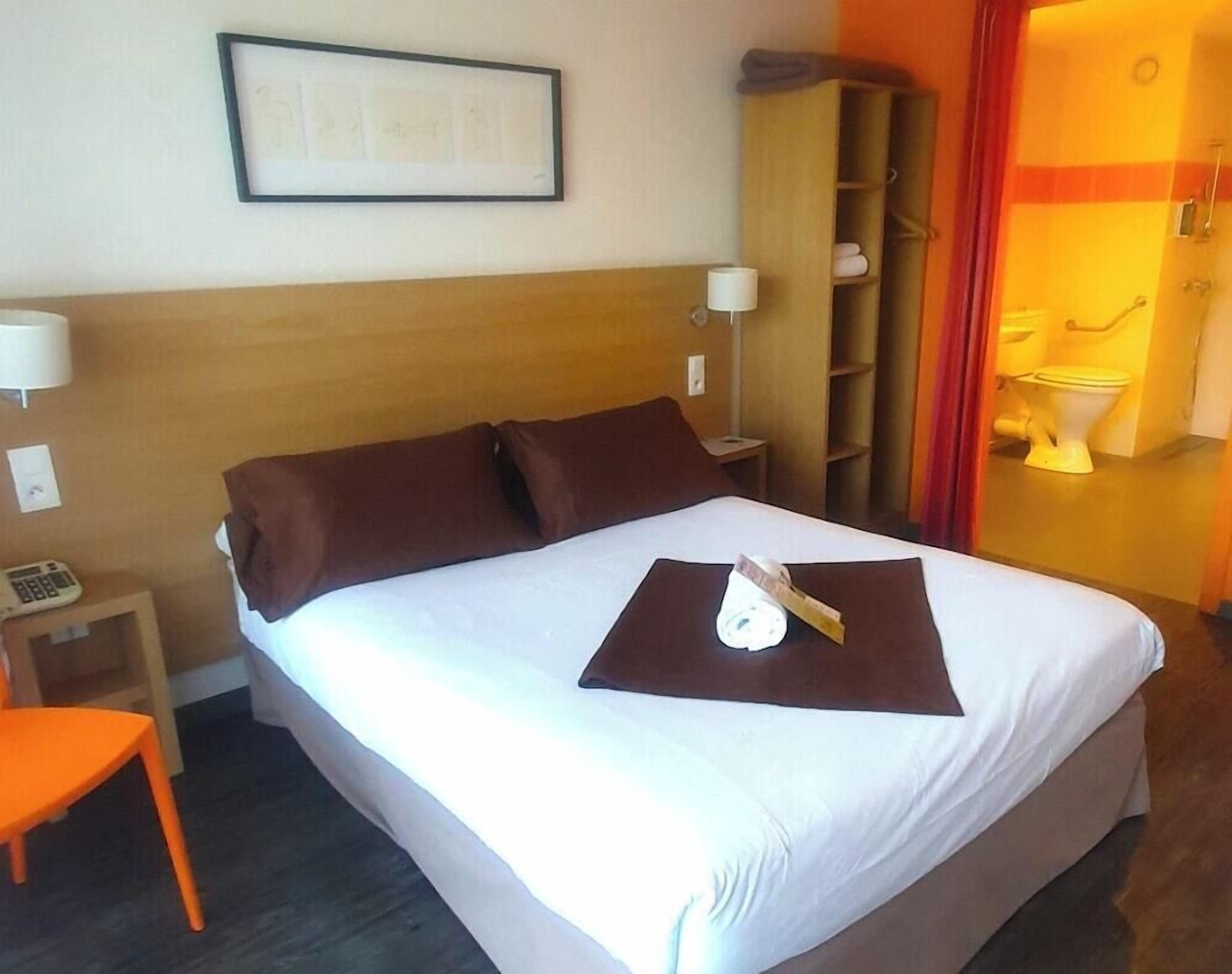 Ein Hotelzimmer mit Bett, Nachttisch, Lampe, Ablagefläche und einem durchgehenden Türspalt sichtbaren Badezimmer.