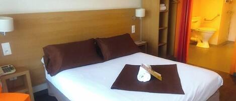 Ein Hotelzimmer mit Bett, Nachttisch, Lampe, Ablagefläche und einem durchgehenden Türspalt sichtbaren Badezimmer.