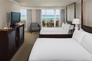 Quarto de hotel com televisão, uma secretária com uma garrafa, uma cadeira, duas camas, uma mesa e vista para o oceano.