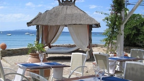 A beachside restaurant with tables and chairs, a thatched-roof structure, and a view of the sea. - Cavaliere Sur Plage (Le Lavandou)