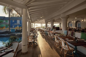 Un restaurante con terraza al aire libre y vistas a una piscina, con pilares blancos y mesas de madera.
