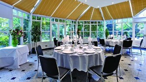 Ein Wintergarten mit runden Esstischen, großen Fenstern und einer hohen Decke mit Holzbalken.