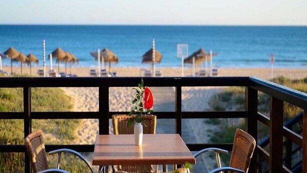 Un restaurante frente a la playa con una mesa y sillas, un jarrón con una flor roja, y sombrillas de playa al fondo.