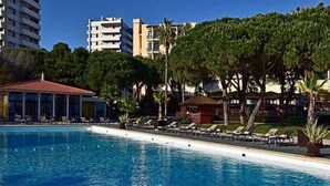 Una piscina con sillas de descanso, un área de estar cubierta y un edificio con balcones al fondo.
