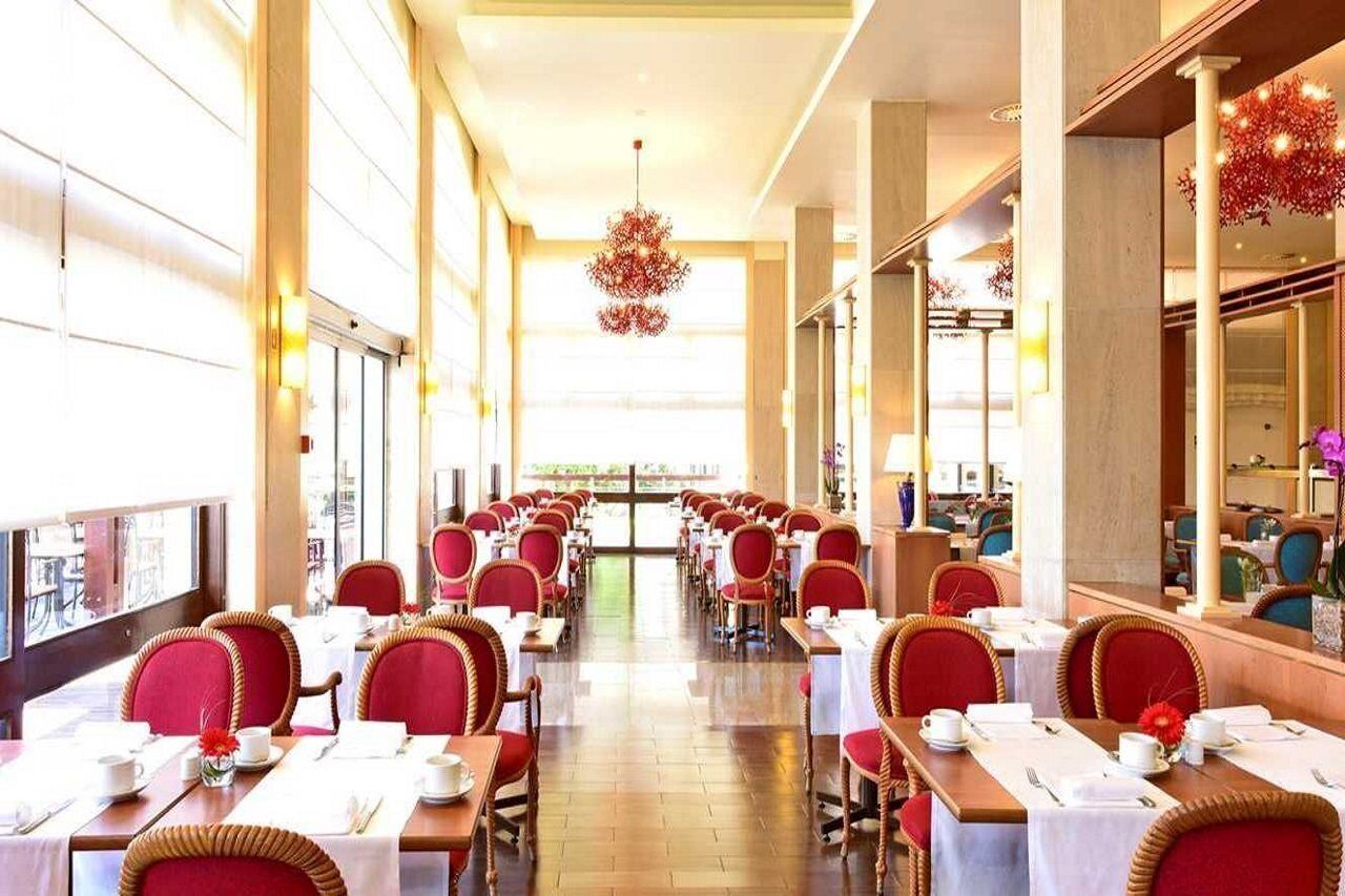 Un restaurante con mesas largas y sillas rojas, ventanas amplias y un candelabro.