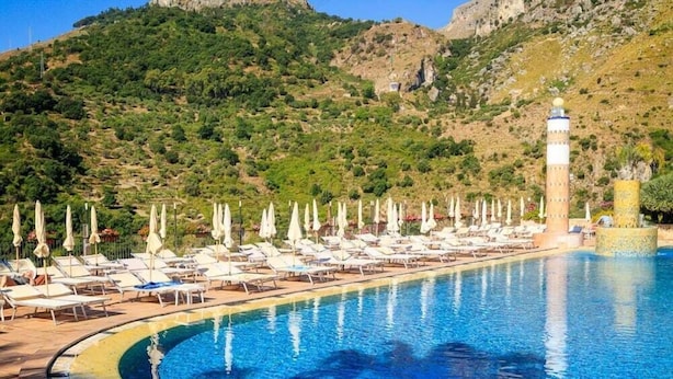 Una piscina con sillas de descanso y sombrillas, rodeada de vegetación y con montañas al fondo.