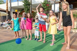 Eine Gruppe von Kindern spielt mit einem Ball auf einer Terrasse, im Hintergrund ist ein Pool und Liegestühle zu sehen.