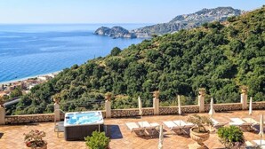 Una terraza en la azotea con hidromasaje, sillones de descanso y plantas en macetas, con vistas a un paisaje costero que incluye el mar y una colina con edificios.