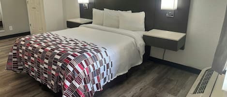 Quarto de hotel com cama, duas mesinhas de cabeceira, um criado-mudo, uma luminária na parede e uma abertura de ventilação.