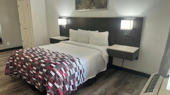 A hotel room with a bed, two bedside tables, a nightstand, a wall-mounted lamp, and a vent.