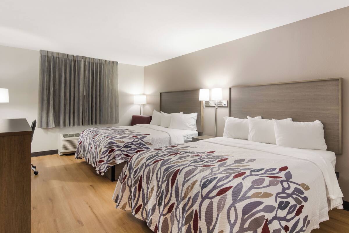 Une chambre d’hôtel avec deux lits, un bureau, une chaise, une fenêtre avec des rideaux et un radiateur fixé au mur.