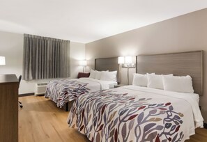 A hotel room with two beds, a desk, a chair, a window with curtains, and a wall-mounted heater.