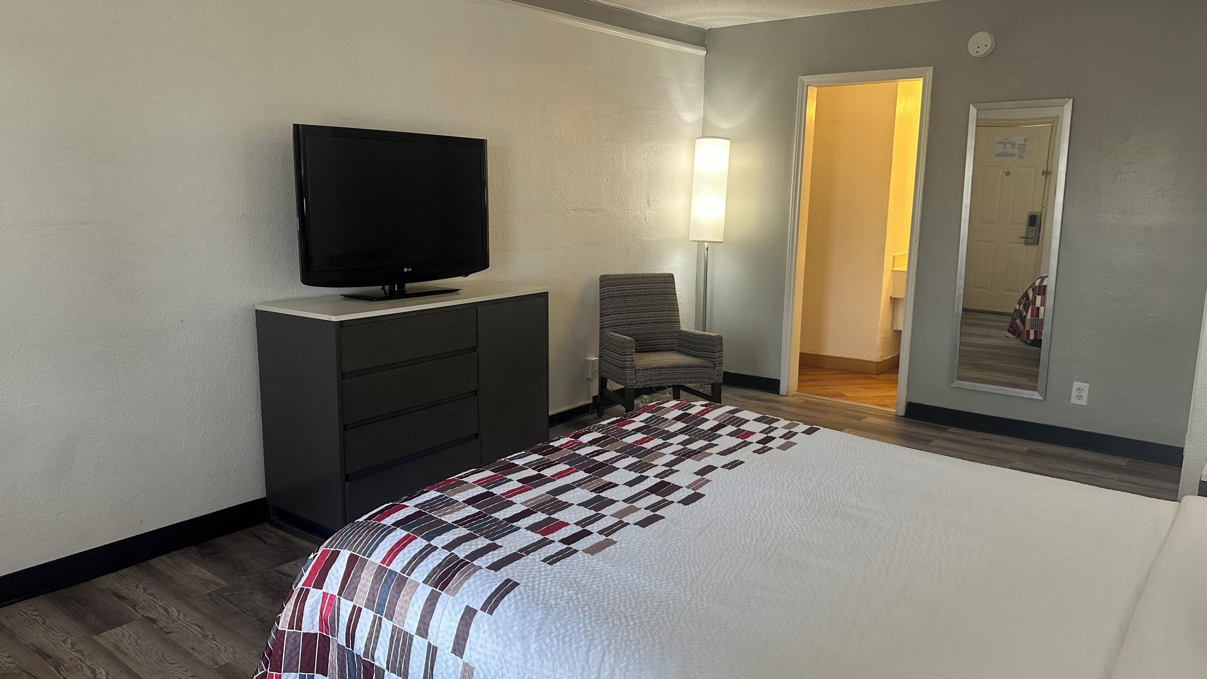 Ein Hotelzimmer mit einem Bett, einem Fernseher auf einer Kommode, einem Sessel und einer Tür zu einem weiteren Raum.