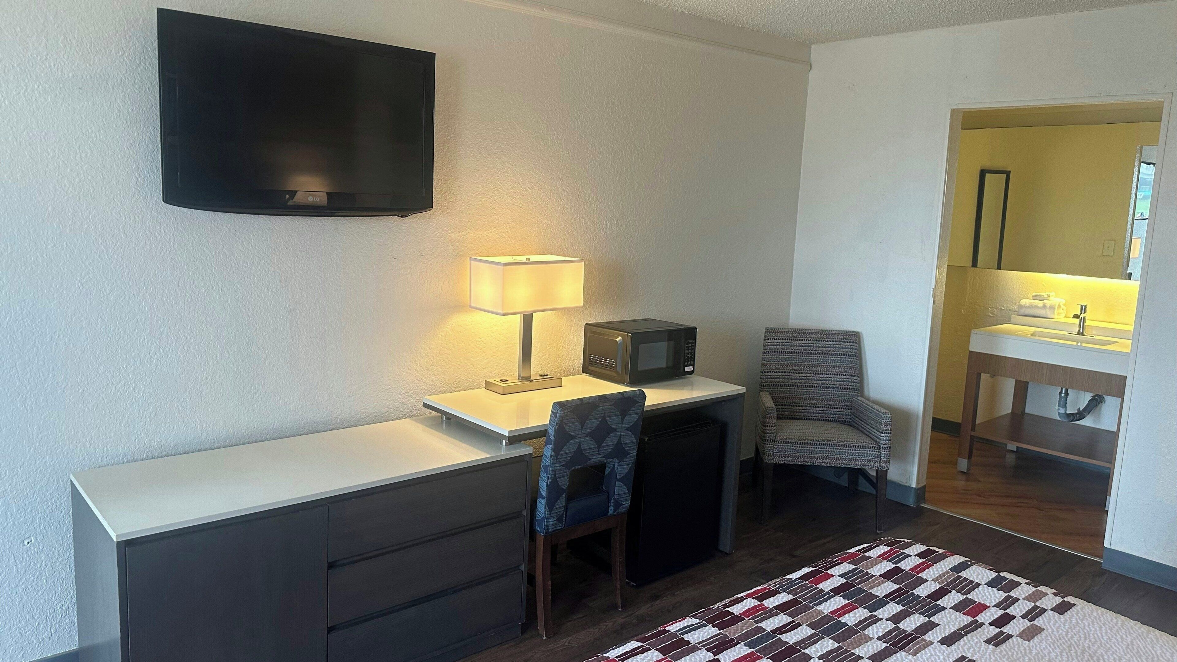 Een compacte kamer met een flatscreen-tv, een bureau met lamp, een magnetron, een stoel en een kleine badkamer met wastafel en spiegel.