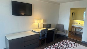 A compact room with a flat-screen TV, a desk with a lamp, a microwave, a chair, and a small bathroom with a sink and mirror.