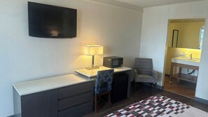 A compact room with a flat-screen TV, a desk with a lamp, a microwave, a chair, and a small bathroom with a sink and mirror.