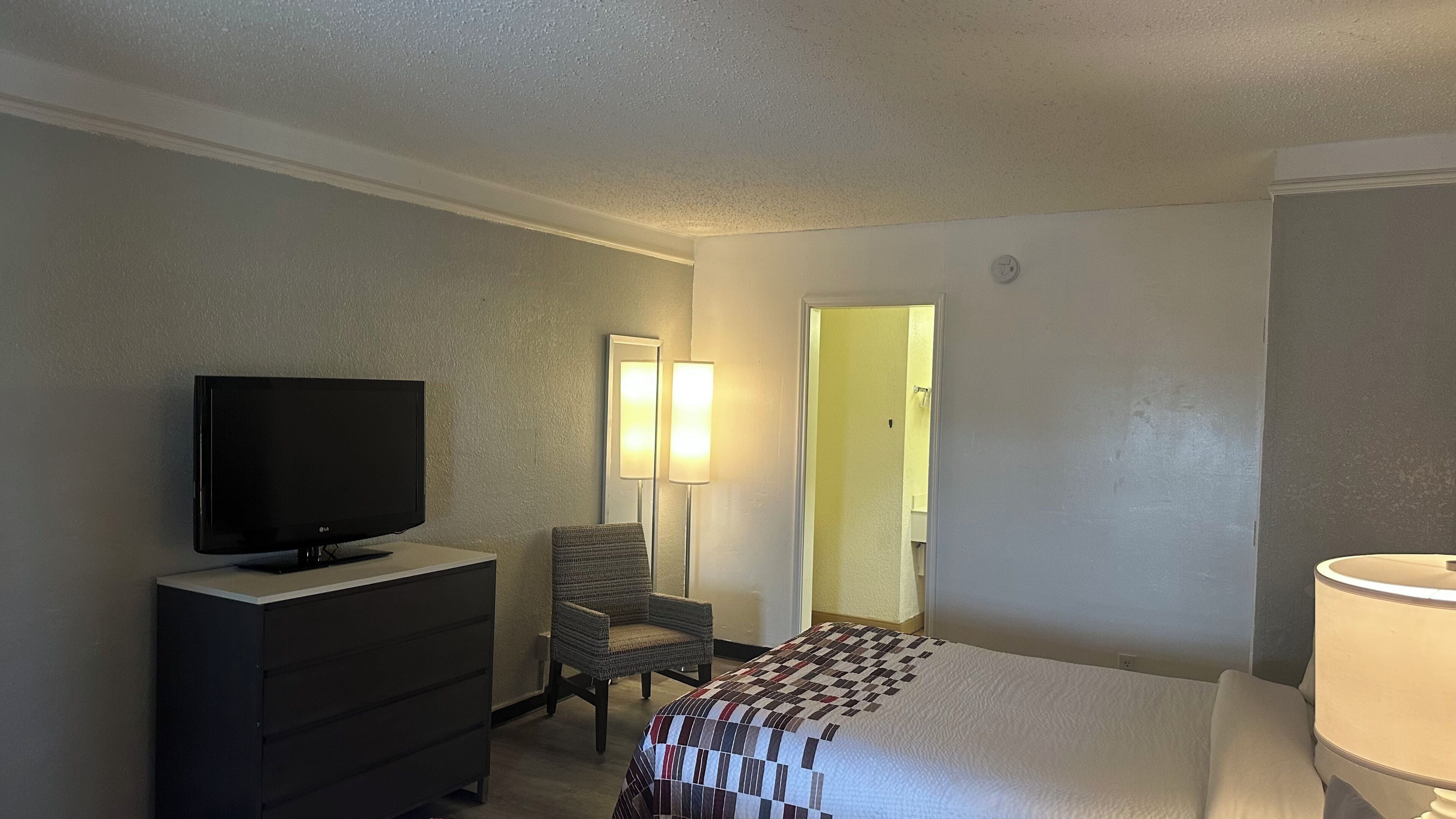 Quarto de hotel com televisão de tela plana sobre um penteadeira, uma cadeira, uma cama com colcha e um abajur.