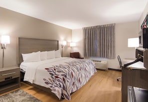A hotel room with a bed, bedside tables, a desk, a chair, a window with curtains, and an air conditioning unit.