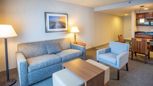 Una sala moderna con sofá, sillón y reposapiés, un comedor con mesa y sillas, y una cocina al fondo.