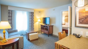 Habitación de hotel con sofá, una mesita con lámpara, televisión sobre un mueble de madera, mesa de comedor con sillas y cama al fondo.