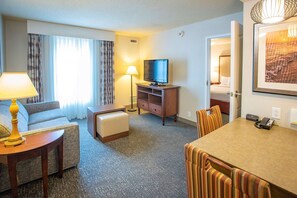 Ein Hotelzimmer mit einer Couch, einem kleinen Tisch mit Lampe, einem Fernseher auf einem Holzständer, einem Esstisch mit Stühlen und einem Bett, das durch eine offene Tür sichtbar ist.