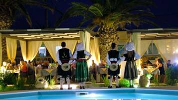 A poolside area with a group of people in traditional attire, a palm tree, and a building with a covered seating area.