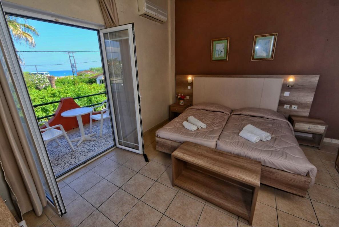 A bedroom with a bed, a bedside table, a nightstand, a wooden bench, a sliding glass door to a balcony with a table and chairs, and two framed pictures on the wall.