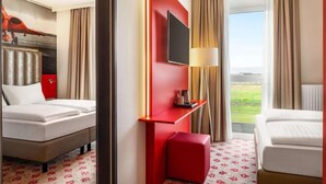 A hotel room with two beds, a TV, a red accent wall, a window with a view, and a red ottoman. - Amedia Airport Graz (Feldkirchen bei Graz)
