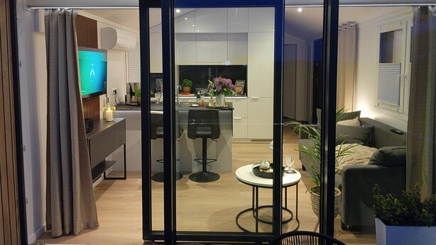 Un intérieur moderne avec une porte vitrée donnant sur une cuisine et un coin repas, un téléviseur à écran plat et un coin salon confortable agrémenté d’une plante.
