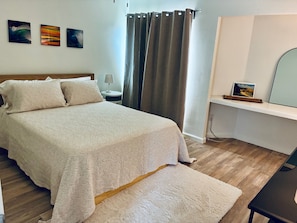 Ein Schlafzimmer mit einem Bett, einem Nachttisch, einer Lampe, einem Schreibtisch, einem Stuhl, einem Fenster mit Vorhängen und Wandbildern.