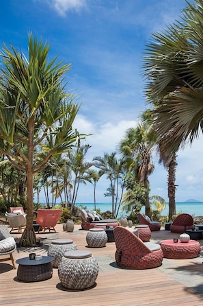 A tropical outdoor lounge area with wicker furniture and palm trees, overlooking a beach. - LUX Grand Gaube Resort and Villa (Grand Gaube)