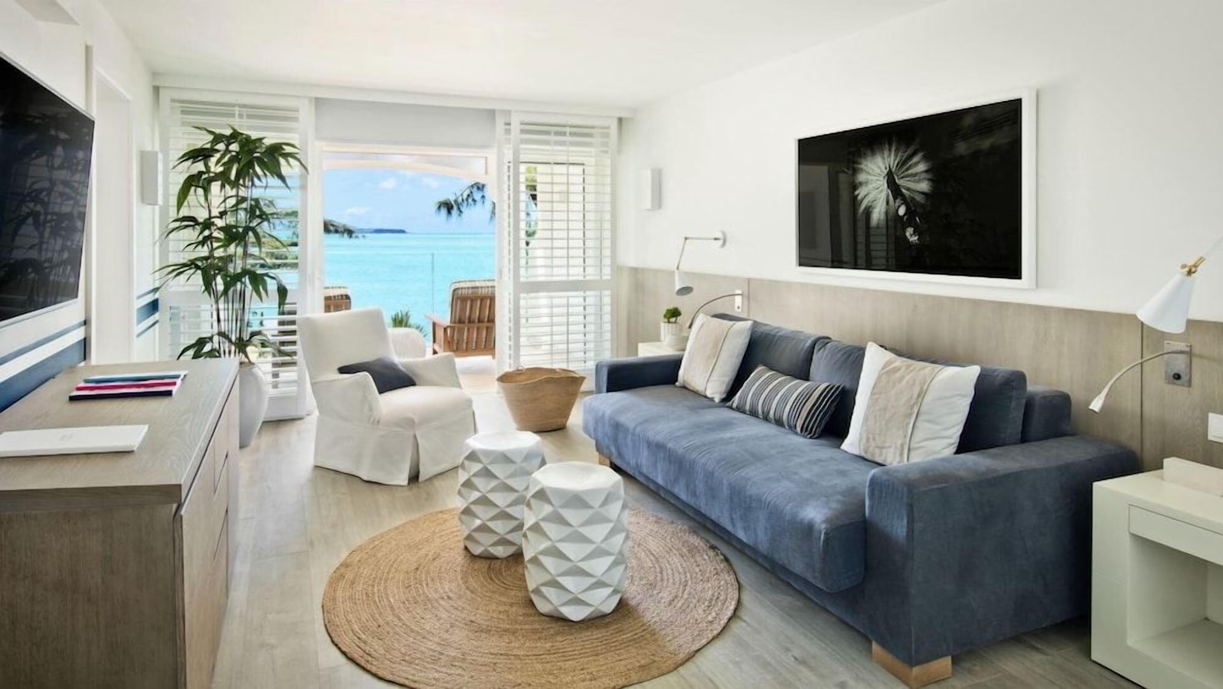 A modern living room with a blue sofa, white armchairs, and a large window overlooking the sea.
