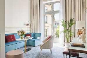 A modern living room with a blue sofa, a round table, a chair, and a large window with curtains. - Hotel Du Louvre (Paris)
