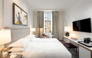 A hotel room with two beds, a desk, a chair, a TV, and a window with a city view. - Hotel Du Louvre (Paris)
