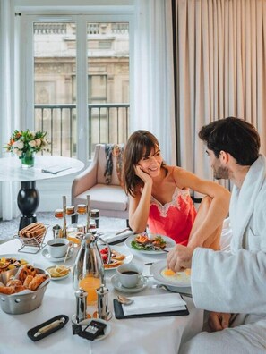 A dining area with a table set for two, a vase of flowers, and a view of buildings through large windows. - Hotel Du Louvre (Paris)