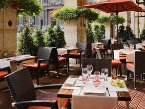 An outdoor dining area with wicker chairs and tables, some with umbrellas, and greenery around. - Hotel Du Louvre (Paris)
