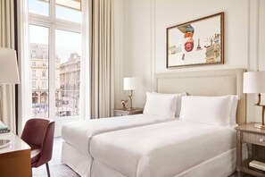 A hotel room with a large bed, a desk, and a chair. There is a window with curtains, a painting on the wall, and a lamp on the bedside table. - Hotel Du Louvre (Paris)
