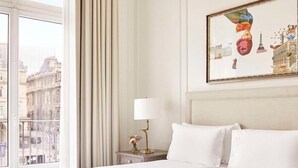 A hotel room with a large bed, a nightstand with a lamp, a painting on the wall, and a view of the city through the window. - Hotel Du Louvre (Paris)