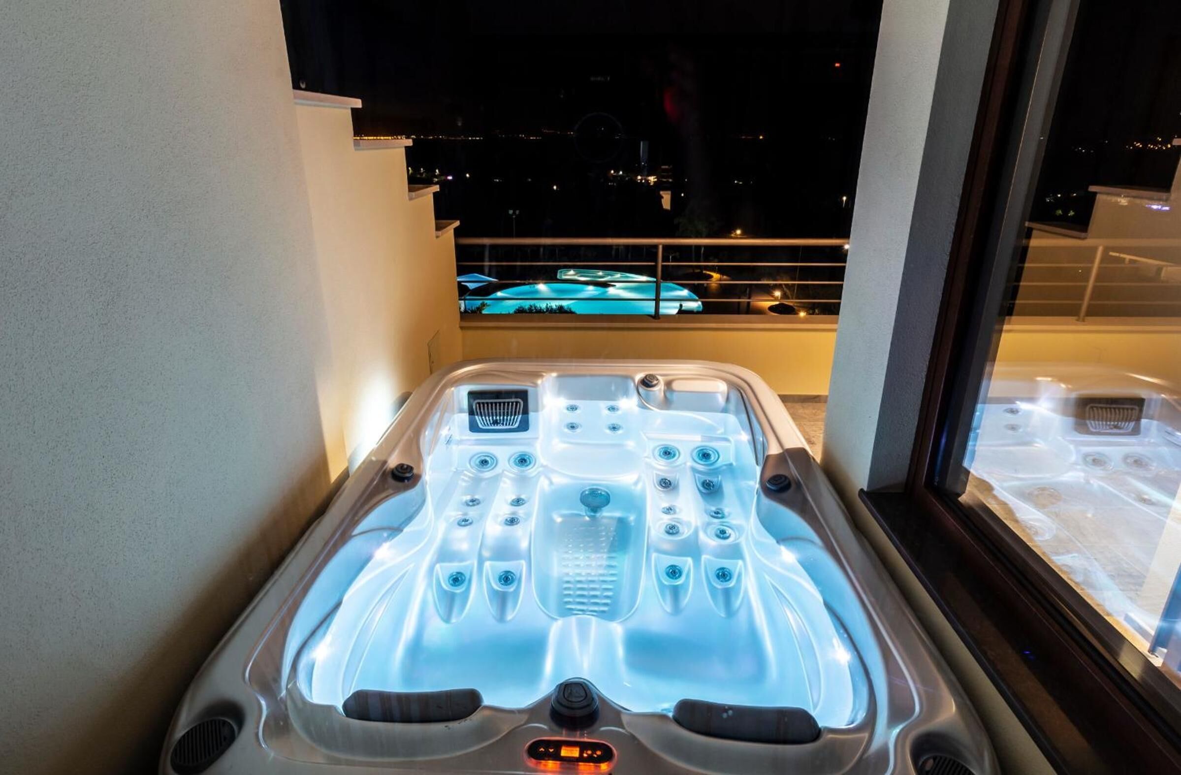 A hot tub with a control panel, a window with a view of a pool and buildings, and a railing.