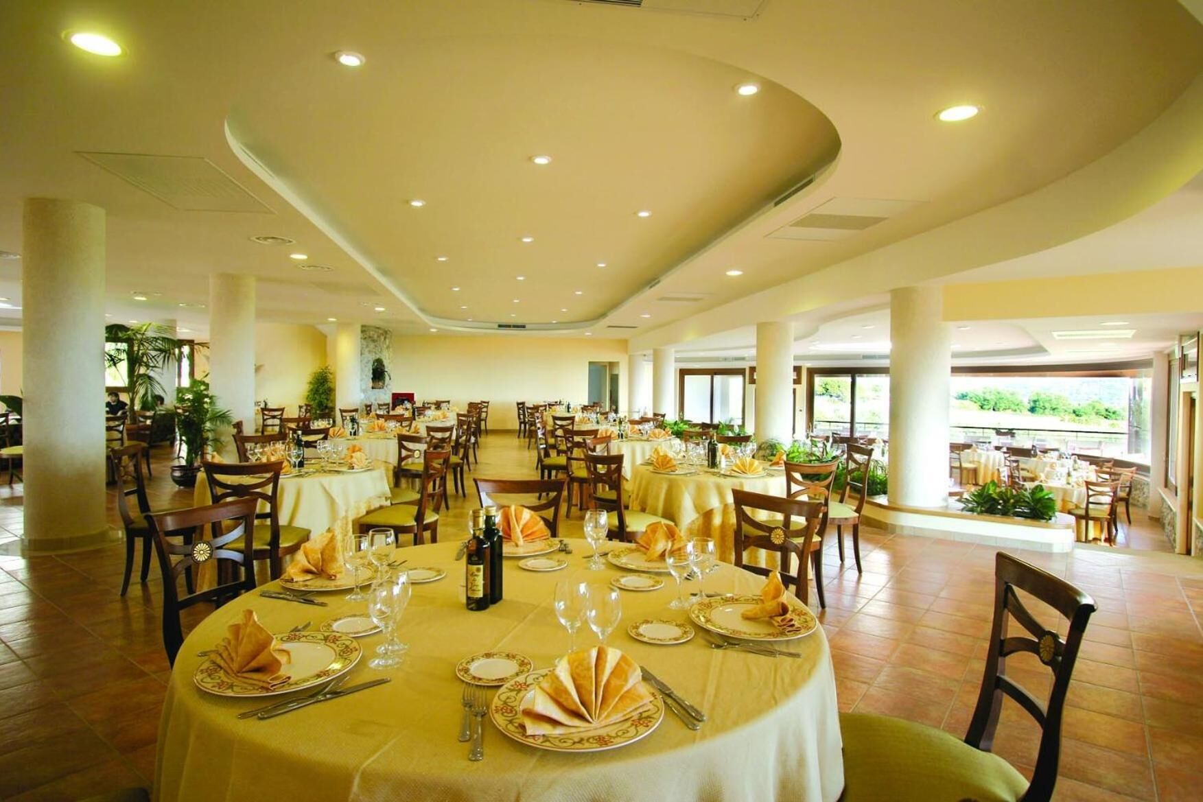 Un'ampia sala da pranzo con tavoli rotondi, sedie e vista sul verde circostante.