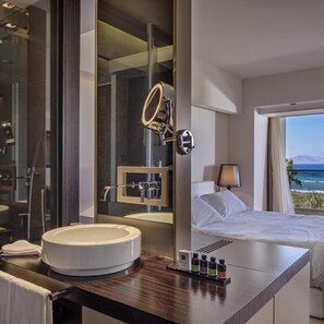 Un hotel moderno con un espejo grande, un lavamanos redondo, una cama con ropa blanca y vista al océano.