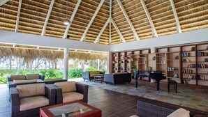 A spacious lounge area with a high thatched ceiling, comfortable seating, a piano, and a large bookshelf. - Portblue Le Sivory Boutique Hotel (Punta Cana)